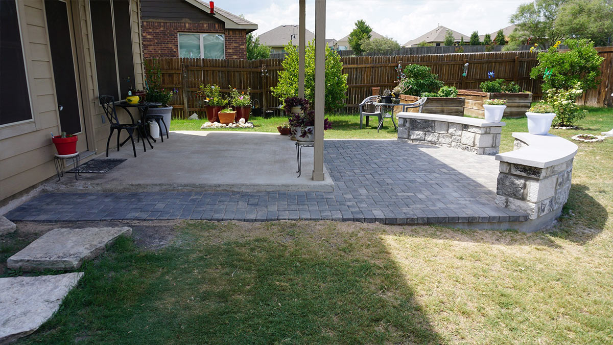 Patio area featuring paver stones, a concrete section, garden planters, and seating against a wooden fence in a sunny backyard.
