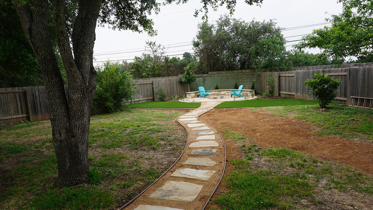 A landscaped backyard featuring a stone path leading to a cozy seating area with turquoise chairs, surrounded by greenery and wooden fences.