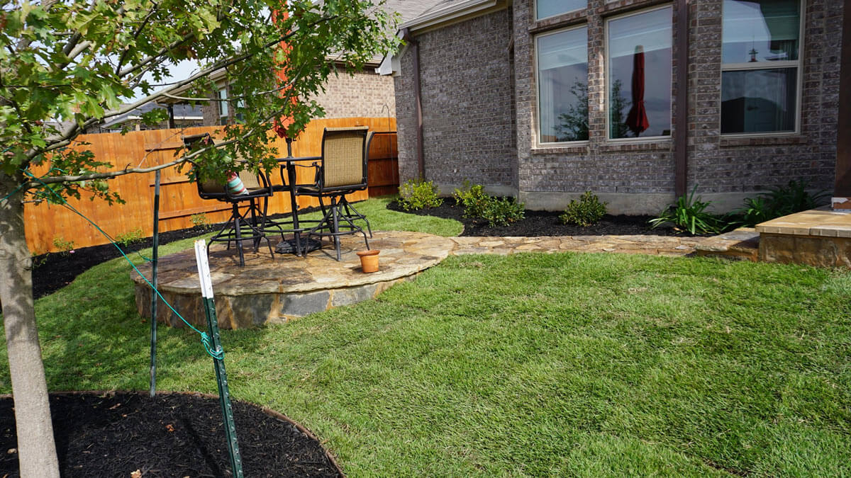 A cozy backyard setting features a stone patio with chairs, lush green grass, a young tree, and a wooden fence in the background.
