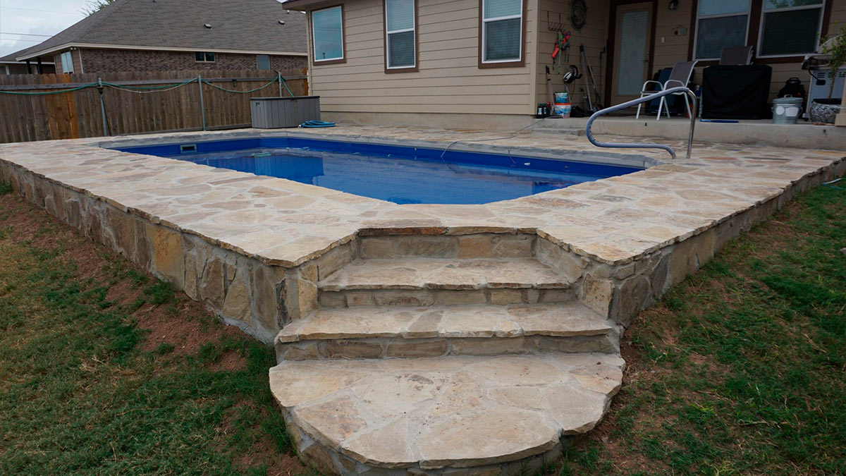 backyard-stone-steps-patio A backyard scene featuring a rectangular swimming pool surrounded by stone steps and patio, adjacent to a modest home and wood fence.