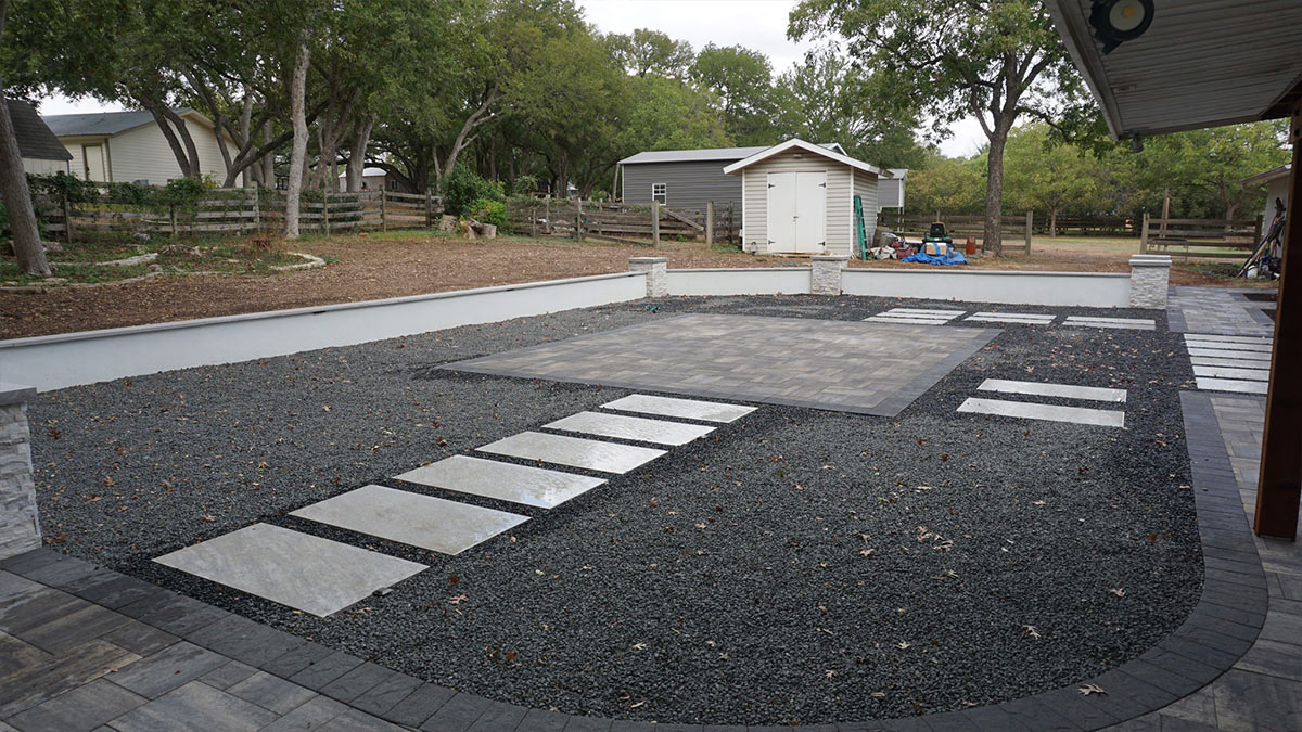 A modern outdoor space features a gravel area with stone pathways, a patio, and surrounding trees, creating a serene backyard environment.
