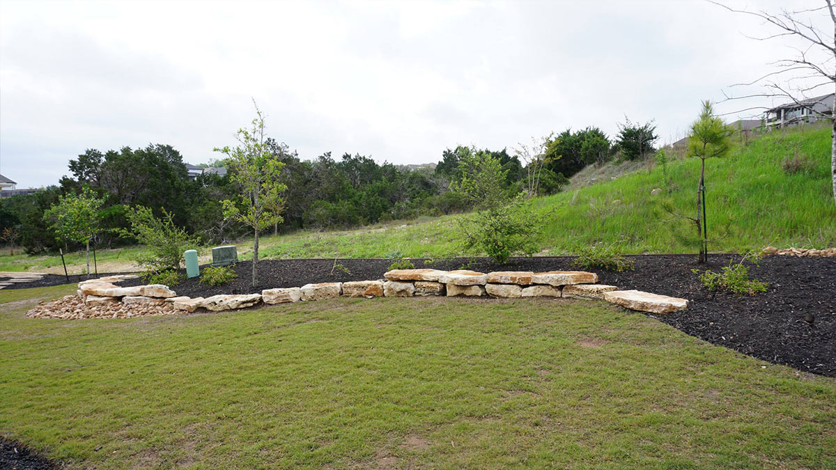 A landscaped garden featuring a curved stone wall, green grass, small trees, and shrubs against a hilly, wooded backdrop.