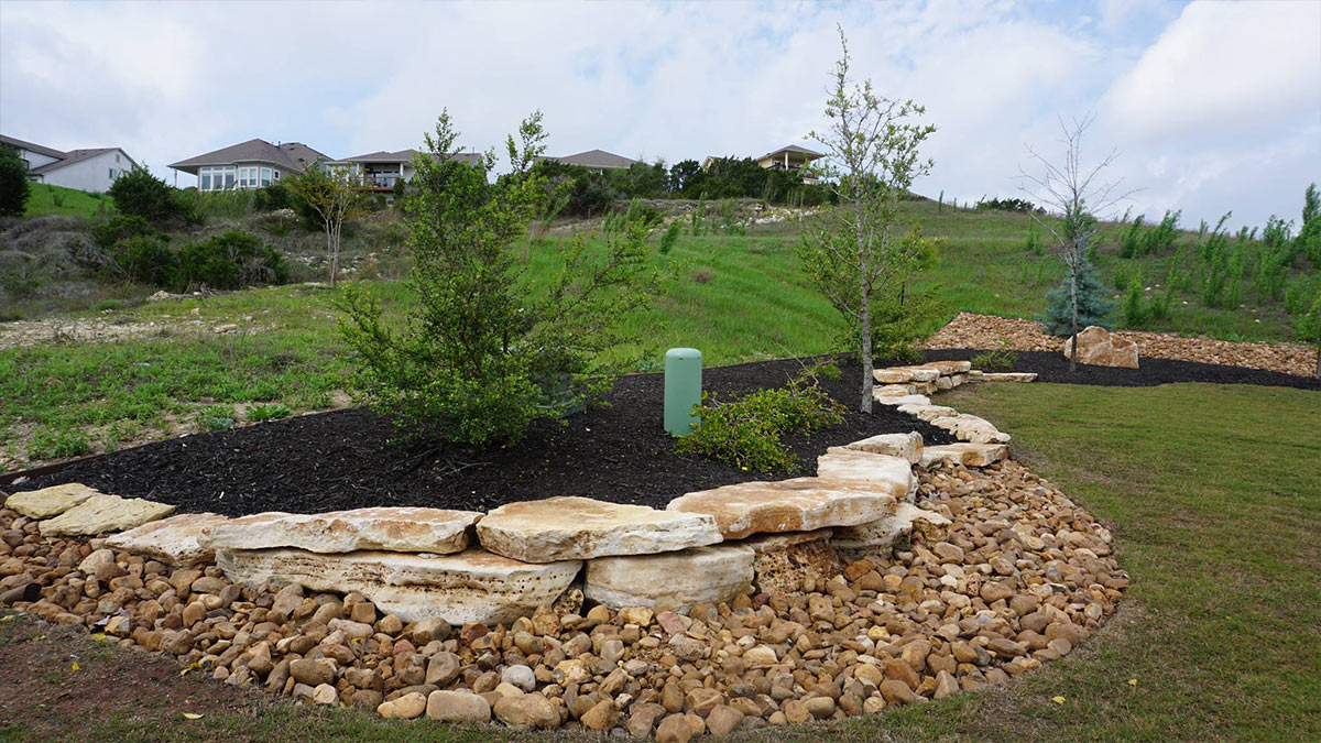 A landscaped garden featuring stone borders, native shrubs, and a grassy hill in the background under a partly cloudy sky.