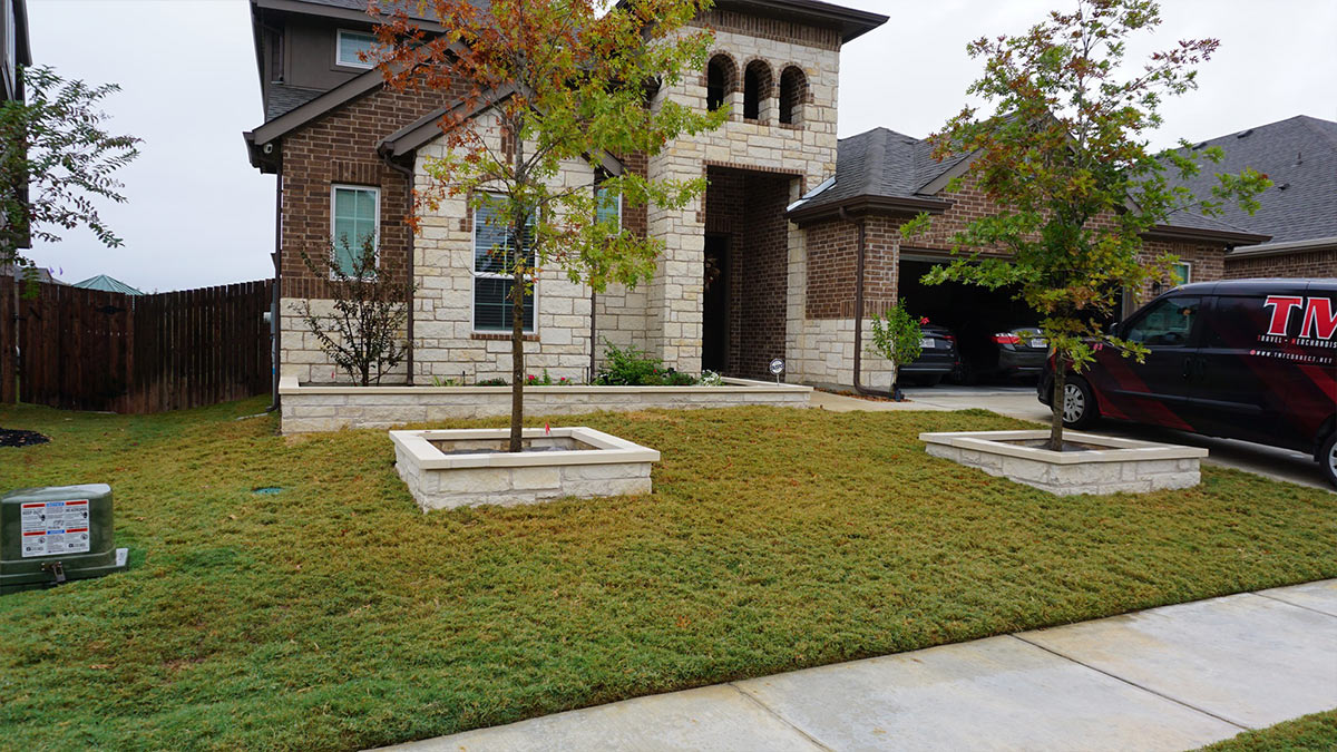 A modern house with stone and brick exterior, featuring landscaped greenery, two trees in planter boxes, and a parked van nearby.