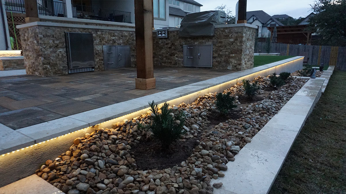 A stylish outdoor area featuring stone walls, a grill station, illuminated landscaping, and lush plants framed by decorative rocks.