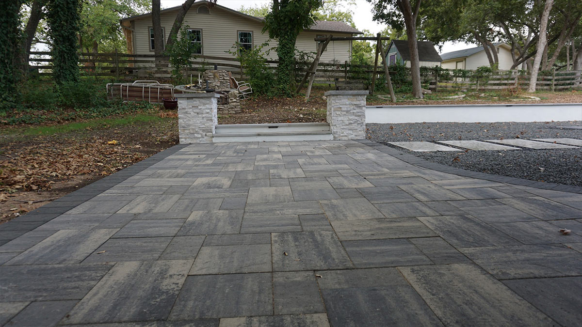 A newly paved outdoor pathway with stone pillars leads to a house, surrounded by trees and a rustic fenced area.