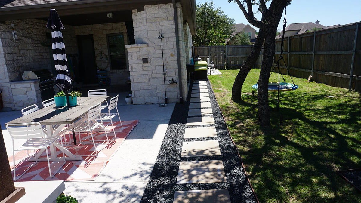 A sunny outdoor patio features a dining table with chairs, striped umbrella, and stepping stones leading to a grassy area with a swing.