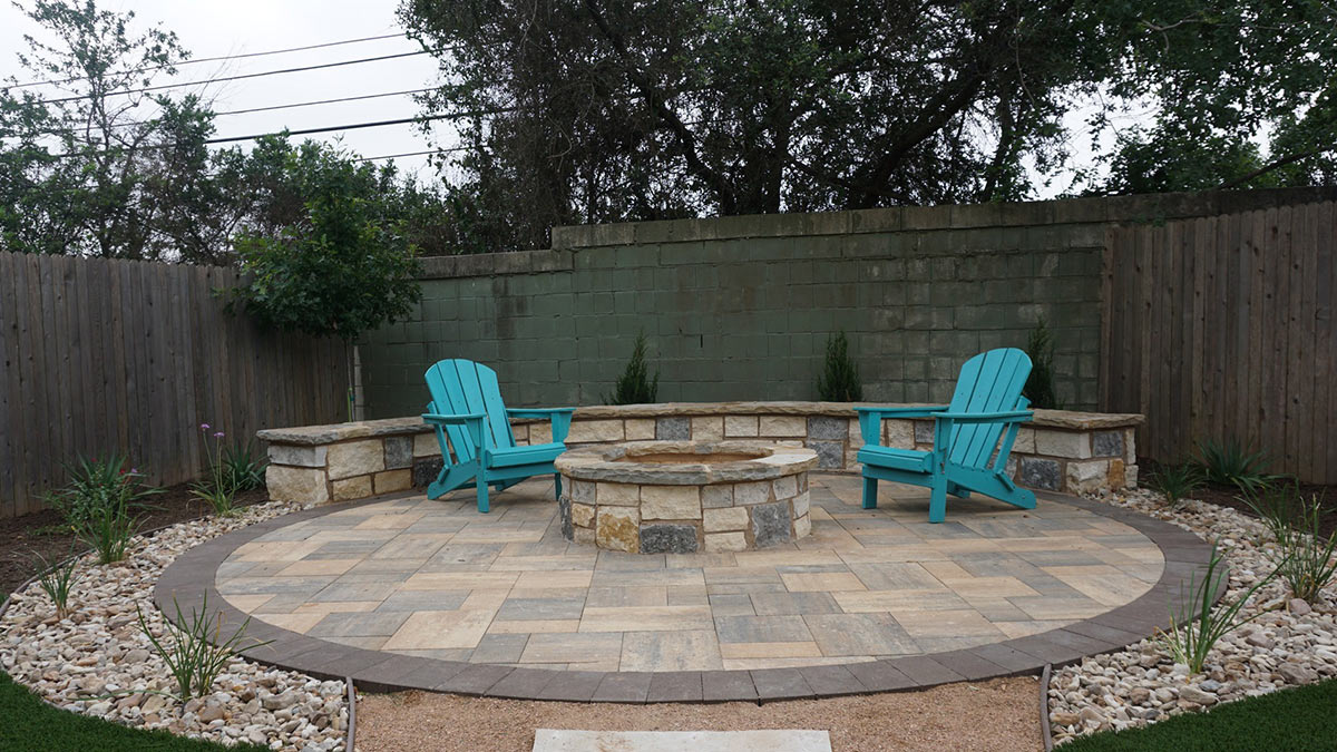 A cozy outdoor patio featuring two turquoise Adirondack chairs and a stone fire pit, surrounded by landscaping and a wooden fence.