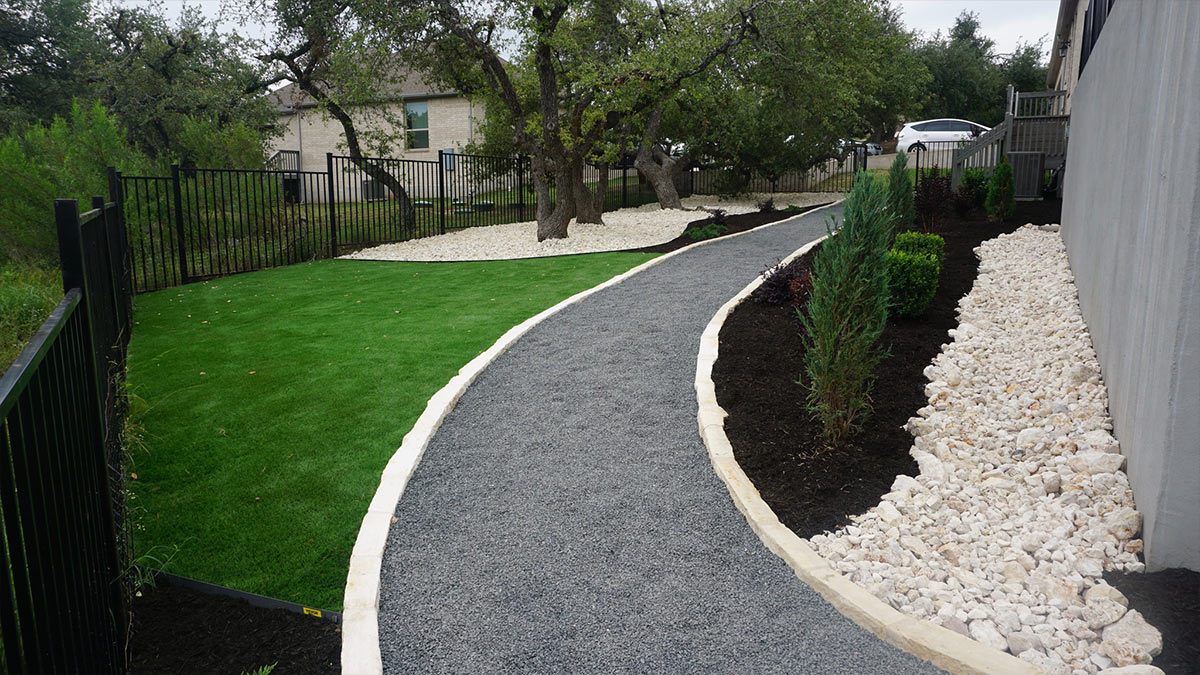 A landscaped pathway curves through green grass and gravel, bordered by decorative rocks and shrubs, next to a fence and house.
