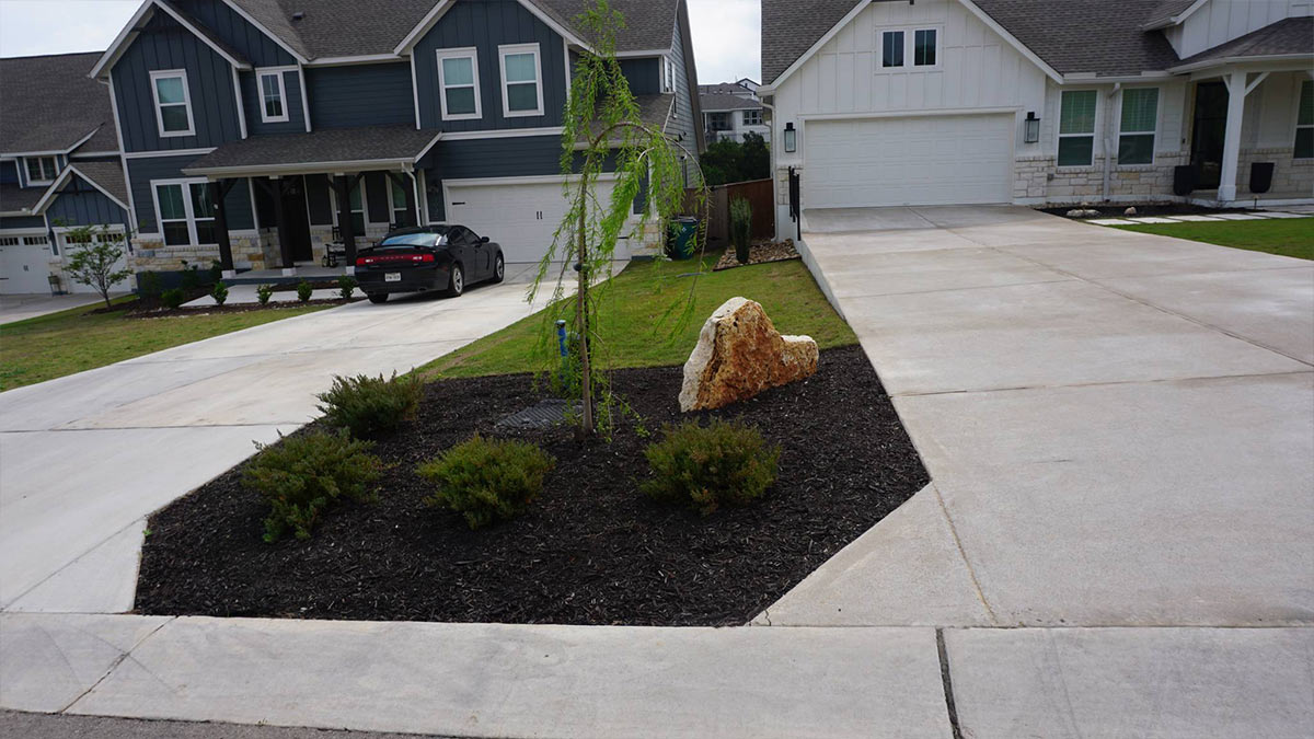paved-driveway A suburban scene featuring two houses, a gray car parked, a small tree, a rock, and a landscaped area with shrubs on a paved driveway.