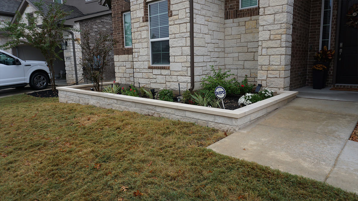 A stone-bordered garden filled with flowers beside a house's entryway, with a parked white truck in the driveway.