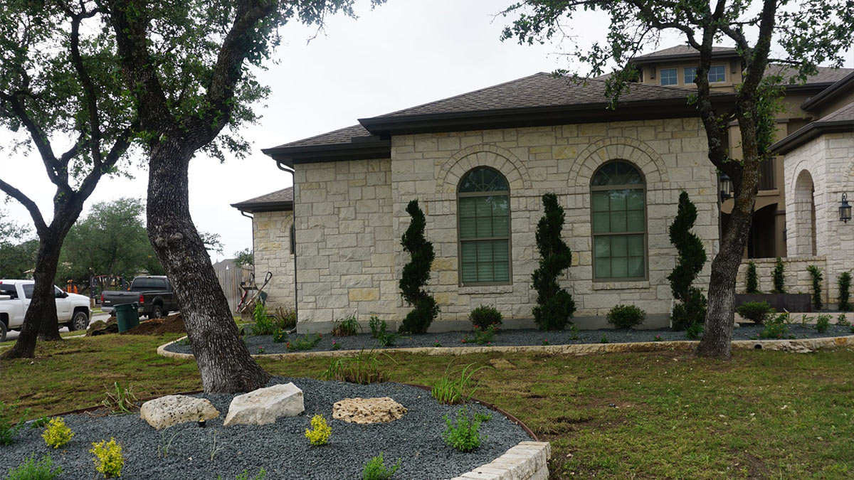 A stone house with arched windows, flanked by manicured shrubs and gravel landscaping, beneath leafy trees on a gray day.