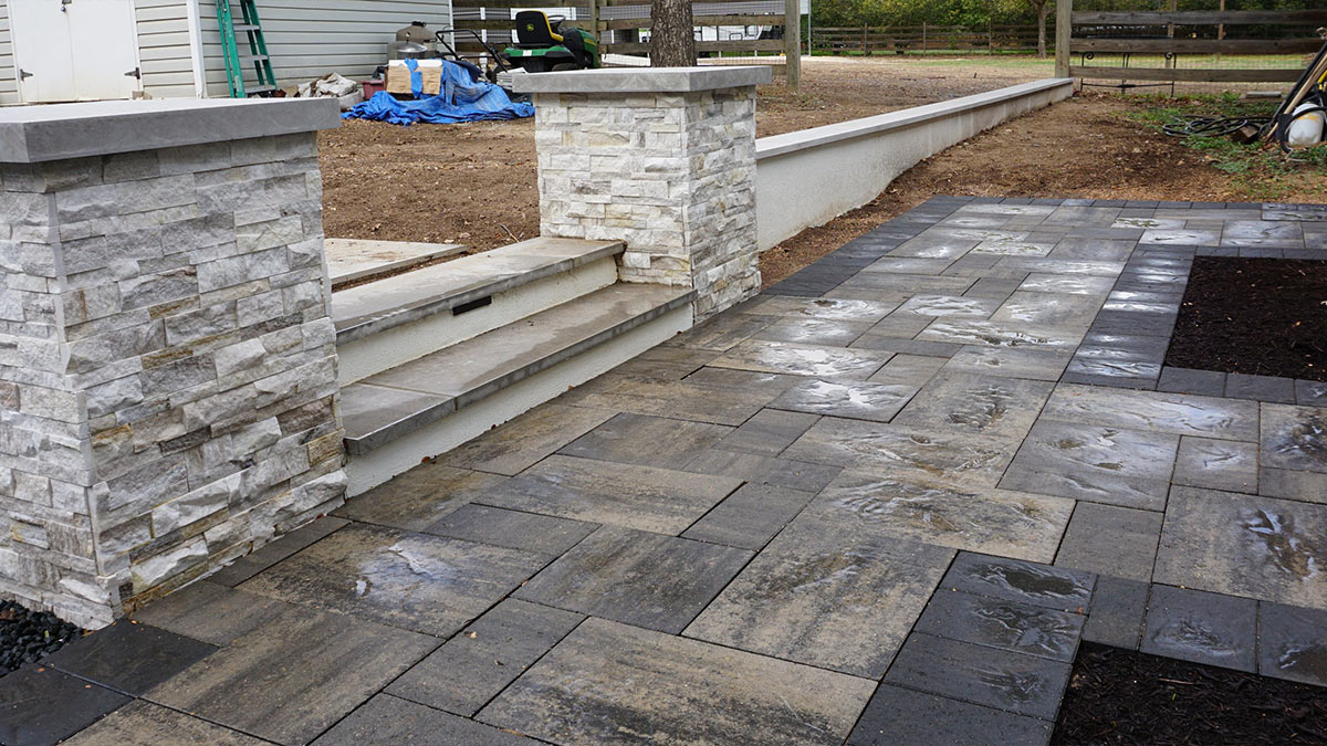 Modern stone steps lead up from a patterned paver patio, framed by two sturdy stone columns, in a freshly landscaped area.