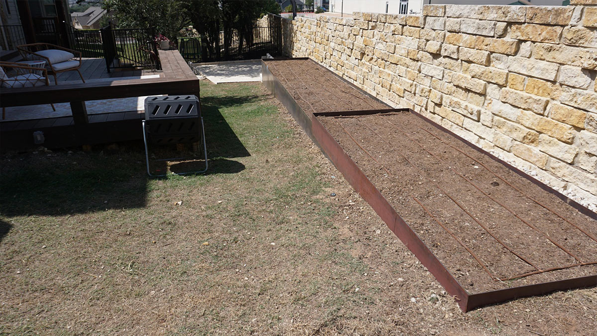 A brown raised garden bed with tilled soil alongside a wooden deck, a compost bin, and a stone wall in a sunny backyard.