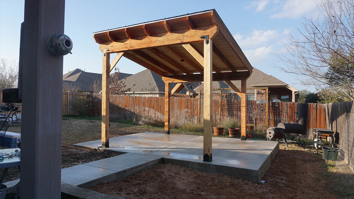wooden-pergola-concrete-slab A wooden pergola with a sloped roof, set on a concrete slab in a backyard, surrounded by a wooden fence and potted plants.