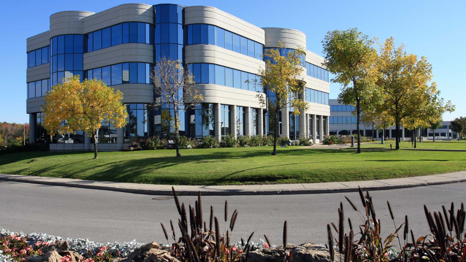 Modern office building with large blue windows, surrounded by manicured lawns and colorful trees under a clear blue sky.