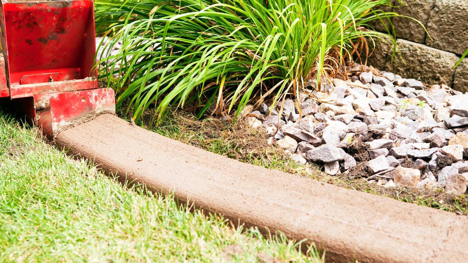 A red concrete mixer laying a smooth concrete strip along a garden edge, beside rocks and green grass.