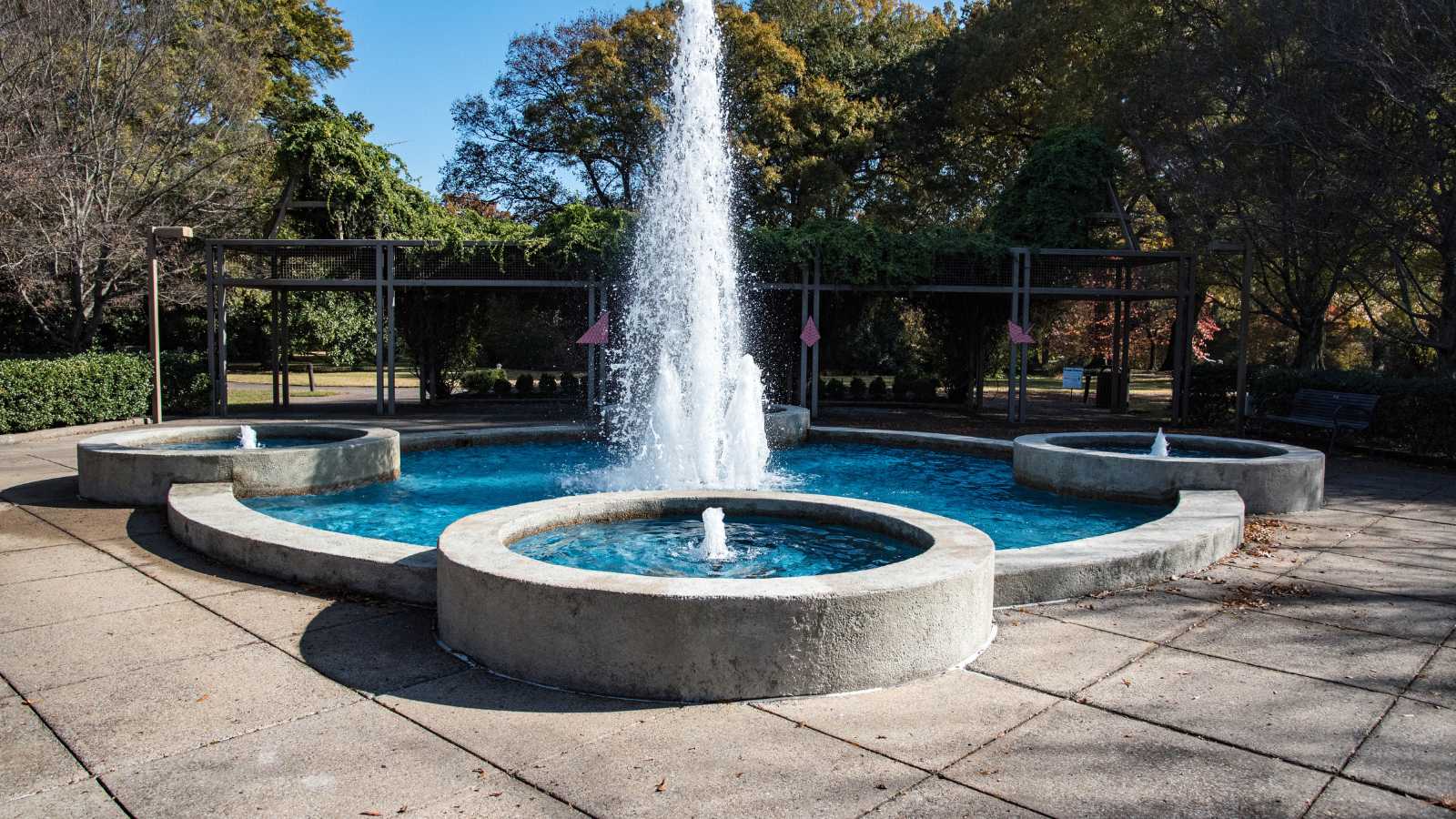 A circular fountain in a park, featuring a central jet of water and three smaller fountains, surrounded by greenery and pathways.
