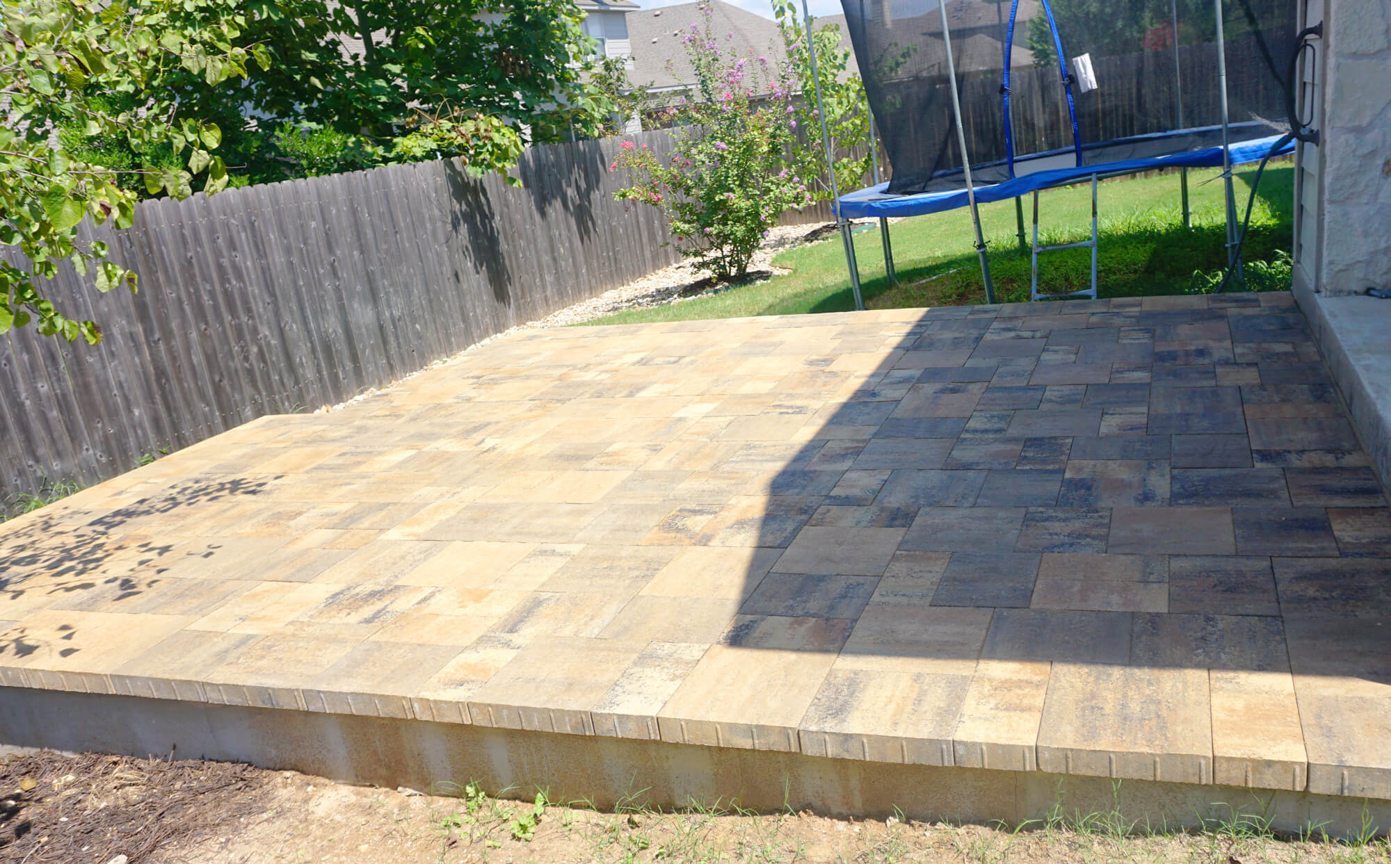 A newly paved patio with rectangular stone tiles, surrounded by grass, a trampoline in the background, and a wooden fence.