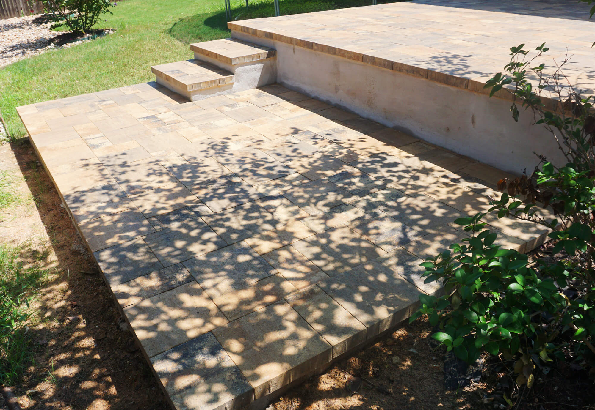 A stone patio with steps, surrounded by greenery, features sunlit tiles casting shadow patterns on the surface.