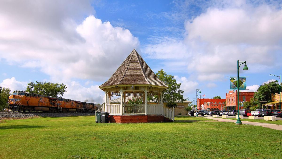 A gazebo in a park with a train passing by in the background, surrounded by trees and green grass.