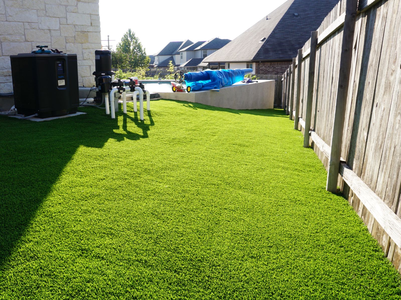 artificial_grass_backyard_landscape A vibrant green artificial grass lawn with nearby equipment and a wooden fence, basking in bright sunlight.