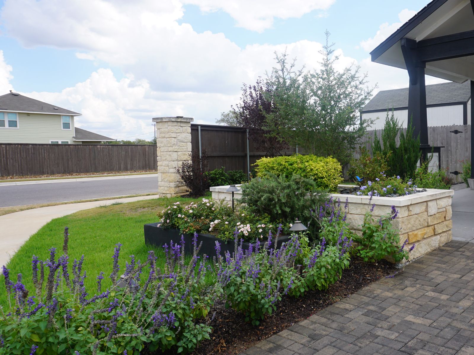 garden_landscape_flowers_shrubs A lush garden with colorful flowers, stone borders, and green shrubs, set against a suburban street and cloudy sky.