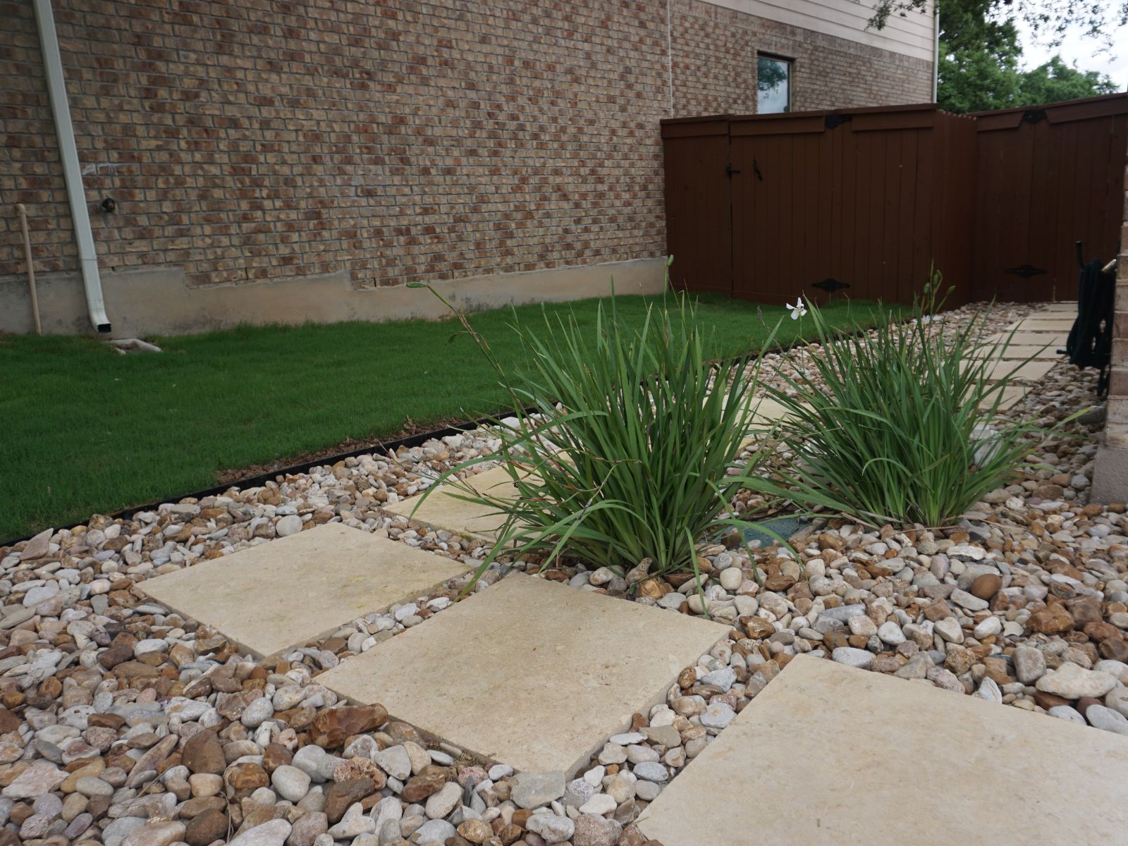 garden_path_stones_grass_plants Garden pathway featuring stone slabs, gravel, and lush green grass, bordered by light brown wooden fencing against a brick wall.