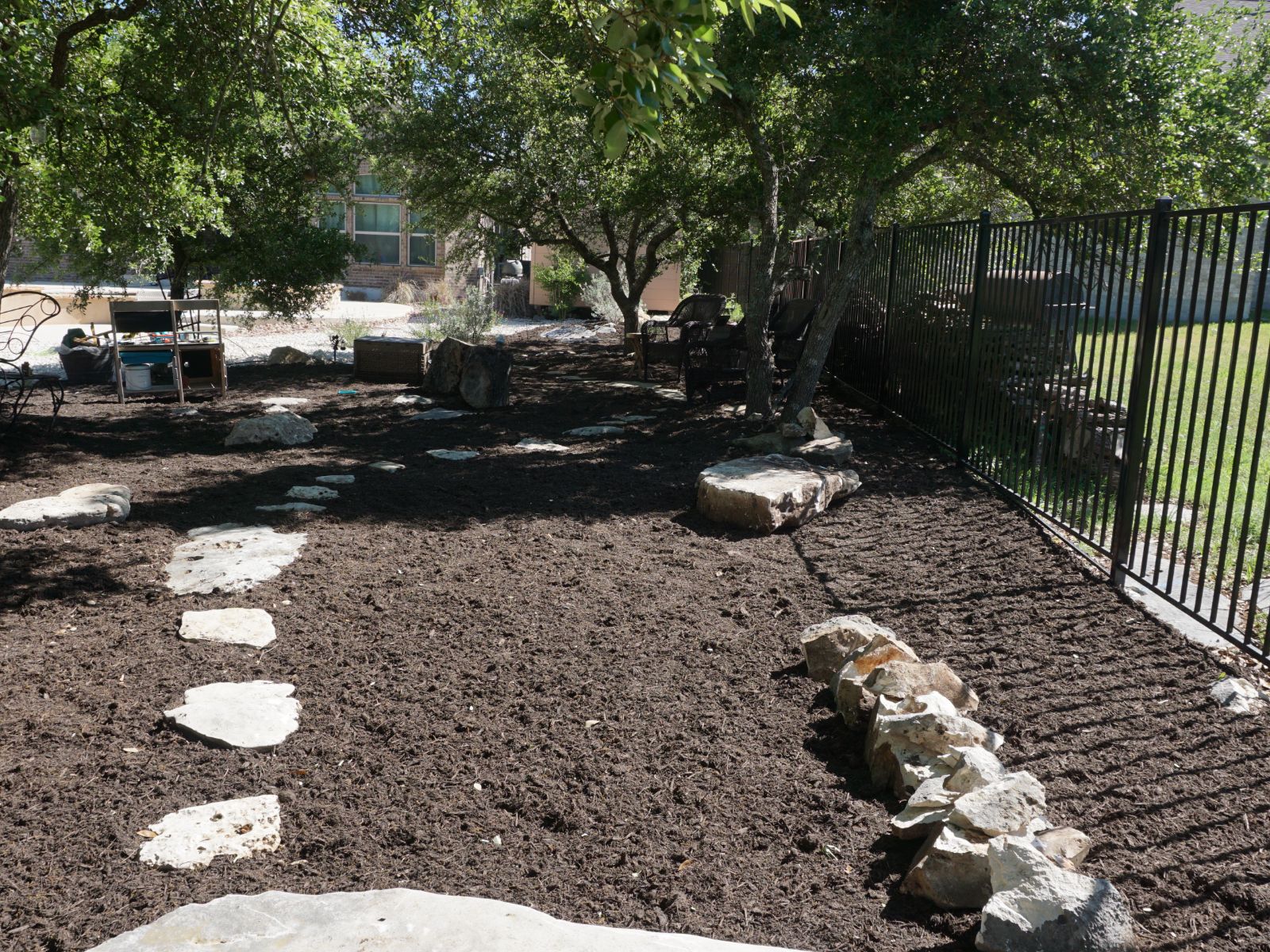 garden_path_stones_mulch A landscaped backyard with stone pathways, mulch, trees, and a black fence, creating a serene outdoor retreat.