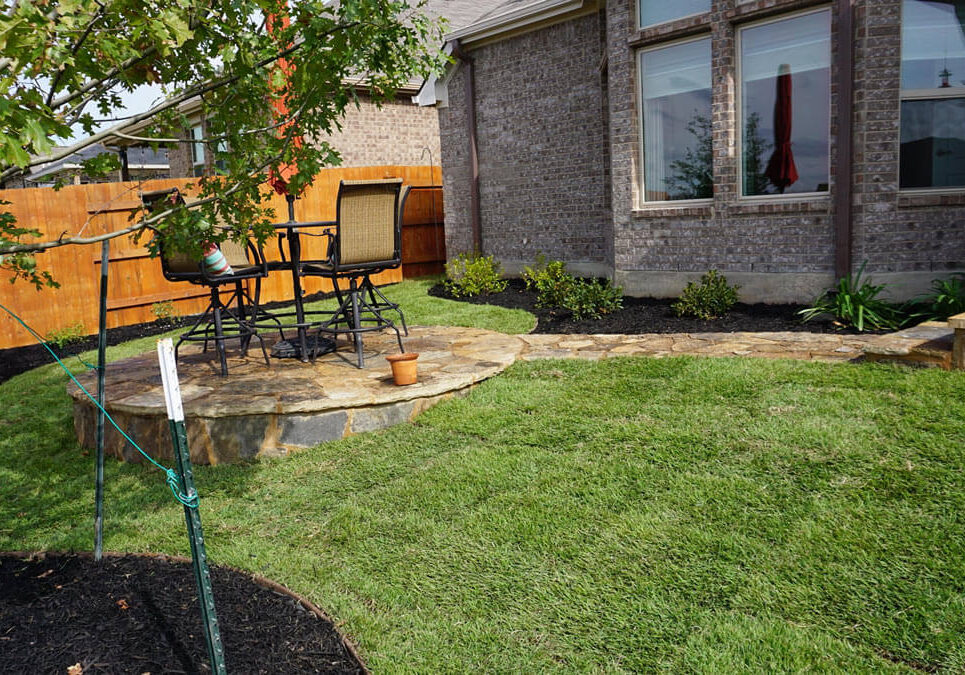 backyard-stone-patio A cozy backyard setting features a stone patio with chairs, lush green grass, a young tree, and a wooden fence in the background.