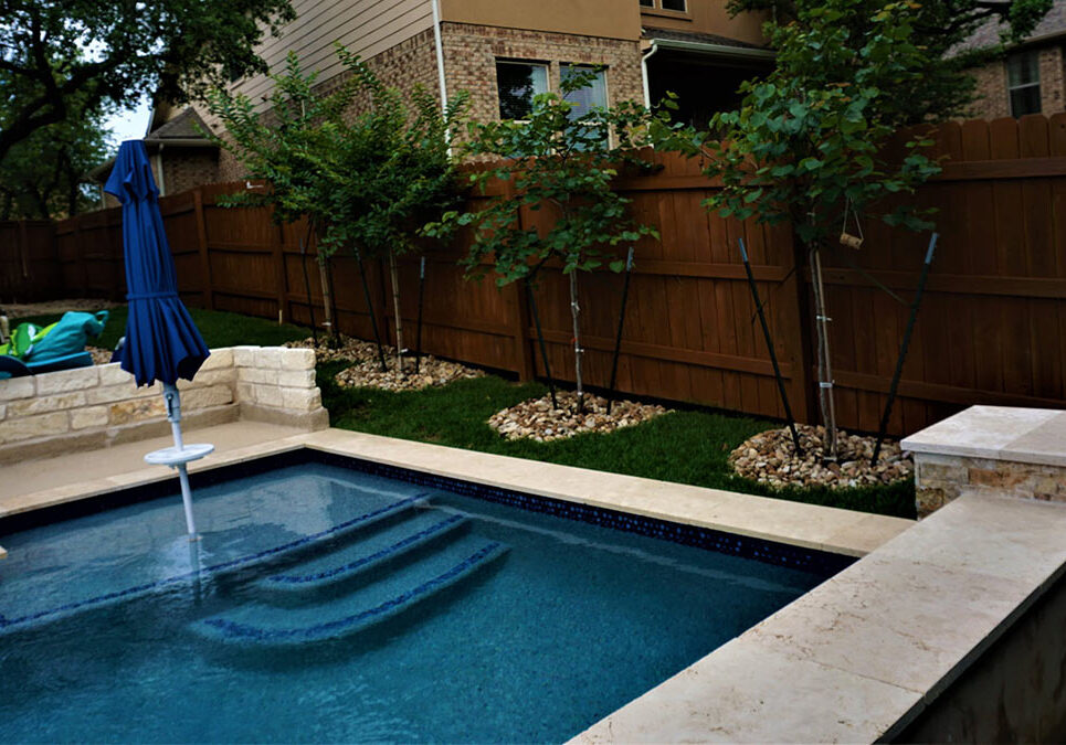 A serene backyard featuring a blue tiled spa with steps, surrounded by green grass and young trees against a wooden fence.