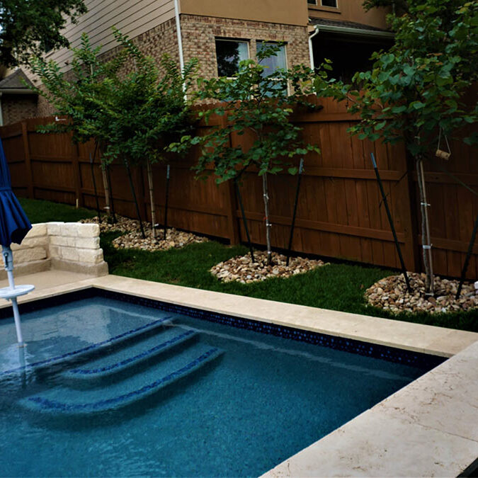 A serene backyard featuring a blue tiled spa with steps, surrounded by green grass and young trees against a wooden fence.