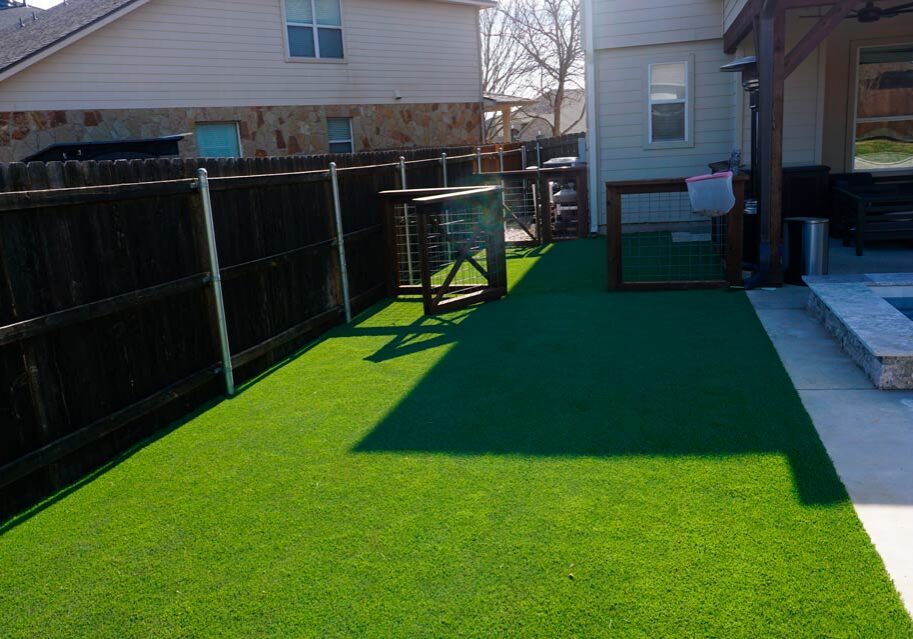 A sunlit backyard with artificial grass, a wooden pet enclosure, and a stone patio with a pool, bordered by a wooden fence.