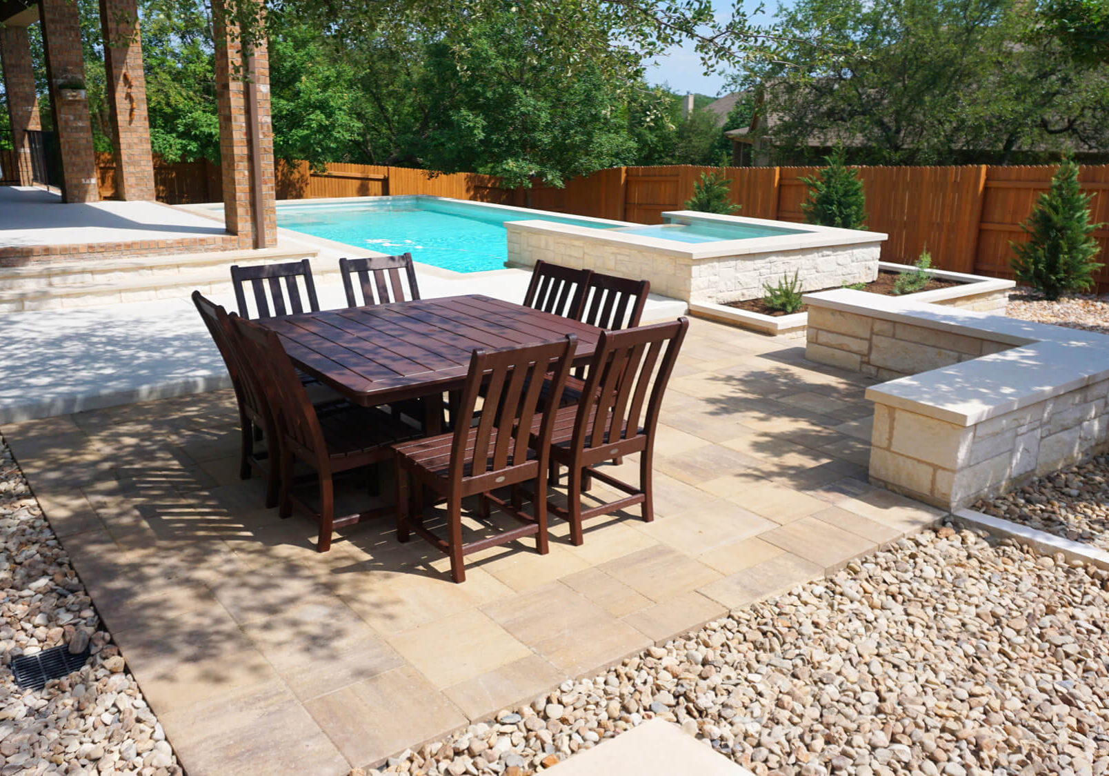 A wooden dining set sits on a stone patio by a swimming pool, surrounded by trees and a wooden fence. Sunlight brightens the outdoor space.