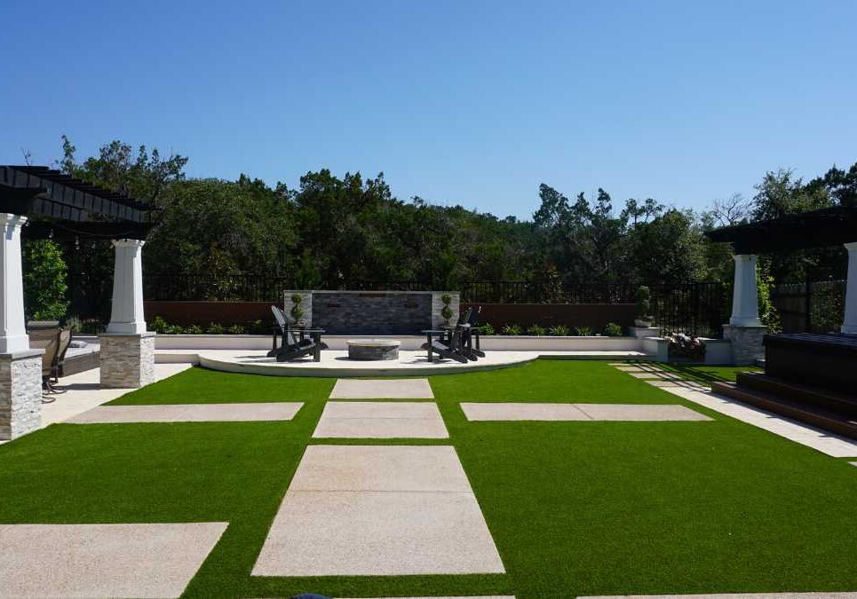 A spacious outdoor patio featuring artificial turf, a fire pit, and shaded seating areas under pergolas, surrounded by greenery and pathways.