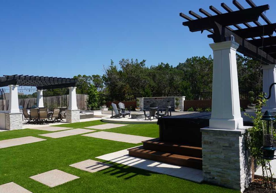 donna before and after8 A spacious backyard featuring a stone patio, stylish pergolas, a hot tub, and lush green grass under a clear blue sky.