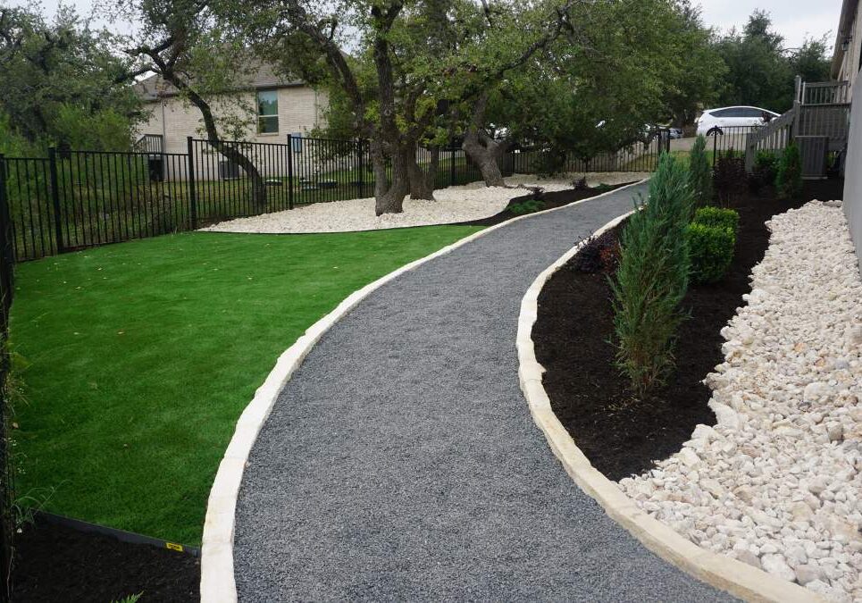 Curved pathway made of gravel and bordered by lush greenery and decorative stones, surrounded by a fenced yard with trees in the background.