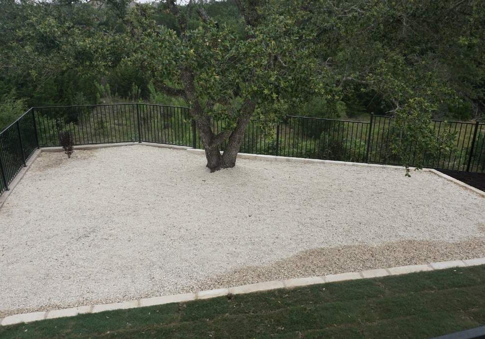 A fenced gravel yard features a lone tree at the center, surrounded by lush greenery in the background.