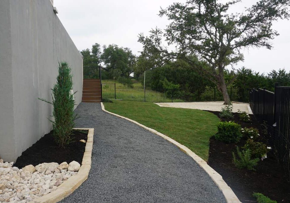 A gravel pathway leads alongside a gray wall, with greenery, plants, and stairs up to a fenced area and grassy hillside beyond.