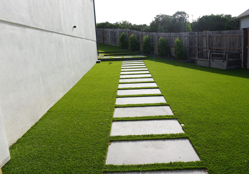 A pathway of concrete stepping stones leads through vibrant artificial grass beside a white wall and wooden fence.
