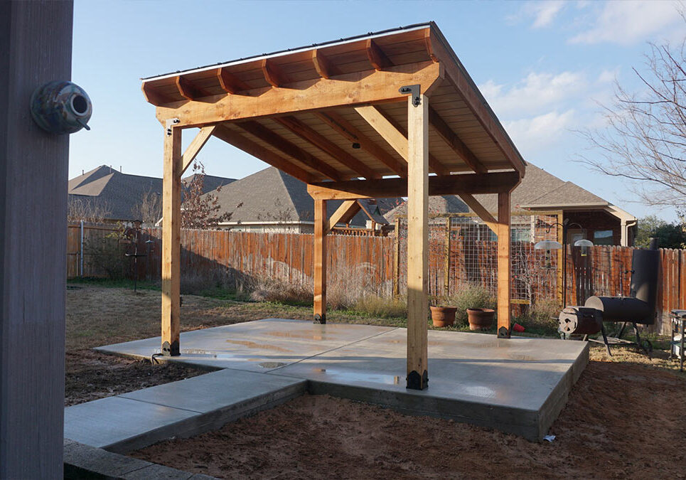 A wooden pergola with a sloped roof, set on a concrete slab in a backyard, surrounded by a wooden fence and potted plants.